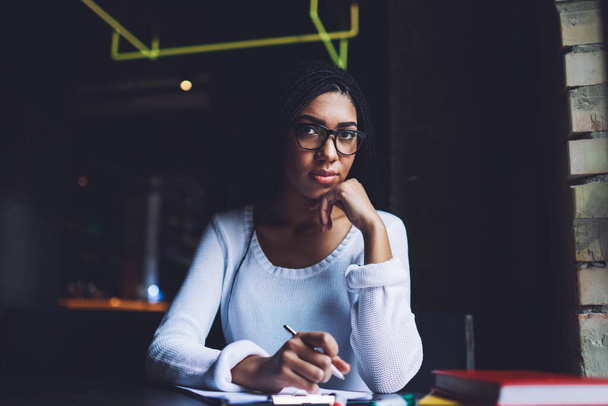 Concentrated African American female freelancer in casual clothes with eyeglasses and hand at chin looking at camera while sitting at table with clipboard - Foto, Bild