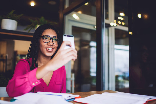 Zadowolony czarny uczennica z warkocze w casual strój i okulary za pomocą smartfona do robienia zdjęć podczas przerwy w odrabianiu prac domowych w kawiarni - Zdjęcie, obraz