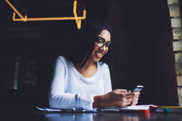 Low angle of happy African American female with toothy smile and eyeglasses sitting at table and browsing mobile phone during break - Фото, изображение