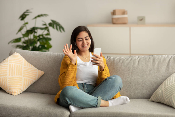 Happy arab lady video calling via cellphone, wearing headphones and waving hand while sitting on sofa at home - Foto, Imagem
