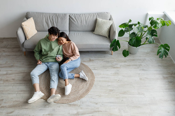 Cheery Asian couple using smartphone application, relaxing on sofa at home on weekend, top view. Copy space - Фото, зображення
