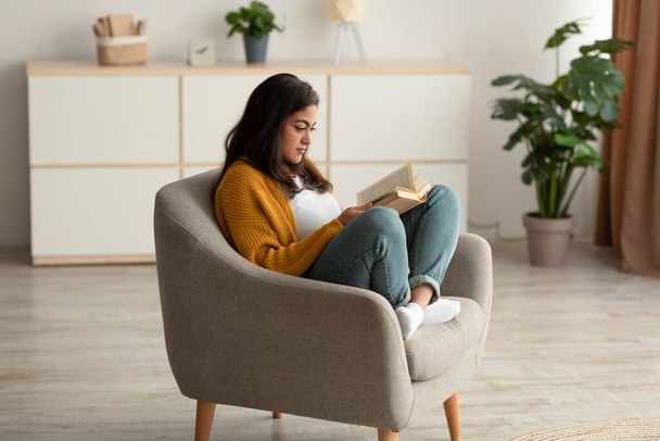 Фокусована арабська леді сидить у кріслі, читає книгу у вітальні, насолоджується лінивим ранком з цікавою історією
 - Фото, зображення