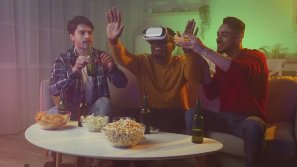 Three multiethnic men friends having fun at home in evening, one guy wearing VR glasses others joking at him, tracking shot - Video
