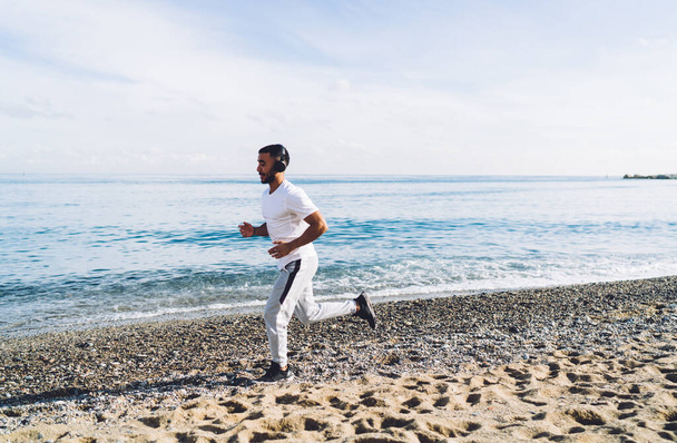 Side view of pensive young ethnic man in sportswear running on sand and listening to music while training on beach - 写真・画像