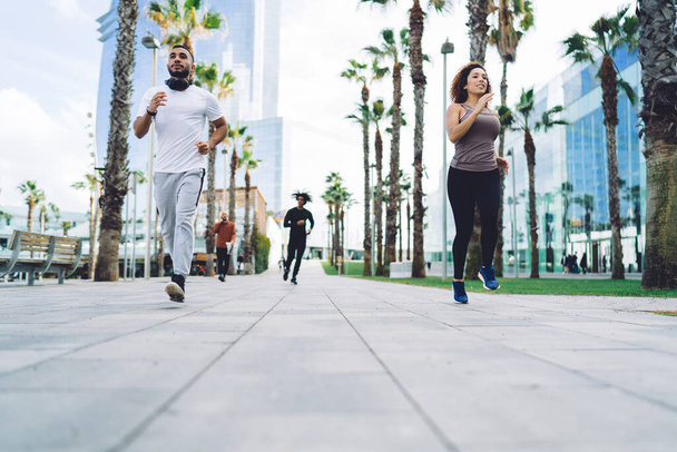 Happy multiracial woman and man enjoying morning jogging in city on street keeping healthy lifestyle, smiling dark skinned female and male in active wear running on urban setting reaching fitness goals - Photo, Image