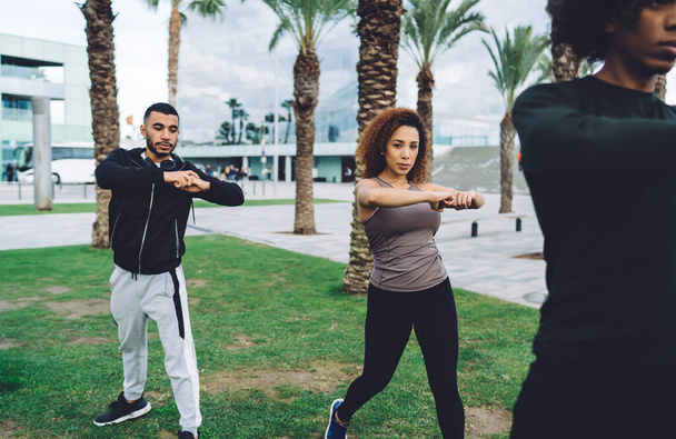 Focused group of multiracial people in sportswear warming up in park on green lawn in city street in summer day - 写真・画像