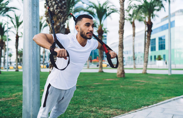 Handsome male in activewear doing exercises on trx equipment motivated with slimming goals, bearded 20s dark skinned sportsman determined training on street having power and energy workout - Fotoğraf, Görsel
