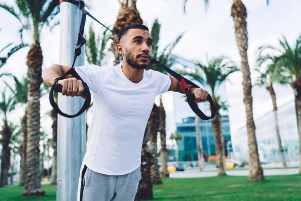 Low angle of confident ethnic bearded male in sportswear doing sport exercise with TRX ropes in park on city street - Foto, imagen
