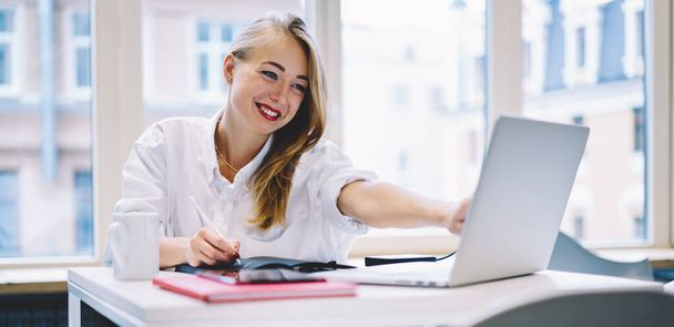 Young positive female in white shirt sitting at table and taking notes while looking and pointing at laptop screen in modern office - Foto, imagen