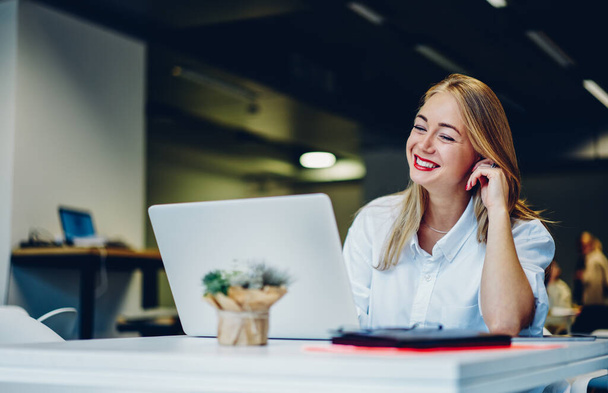 Happy remote worker wearing casual white shirt and red lipstick sitting in modern workplace and using computer at table for work - Foto, imagen
