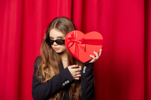 Uzun saçlı, güneş gözlüklü, siyah ceketli, kırmızı perde arkasında kalp şeklinde bir hediye. - Fotoğraf, Görsel
