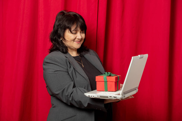 Kırmızı perde arkasında dizüstü bilgisayarı ve hediye kutusu olan 40 yaşında bir kadın. - Fotoğraf, Görsel