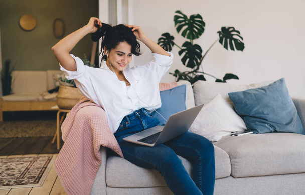 Smiling young female in casual outfit sitting on cozy couch in modern living room while watching movie or having video call on laptop and adjusting hair - Fotografie, Obrázek