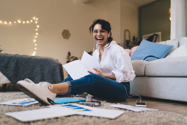 Side view full length delighted young female in casual wear sitting on floor with papers and listening to favorite music via earphones while looking at camera with laughter - 写真・画像