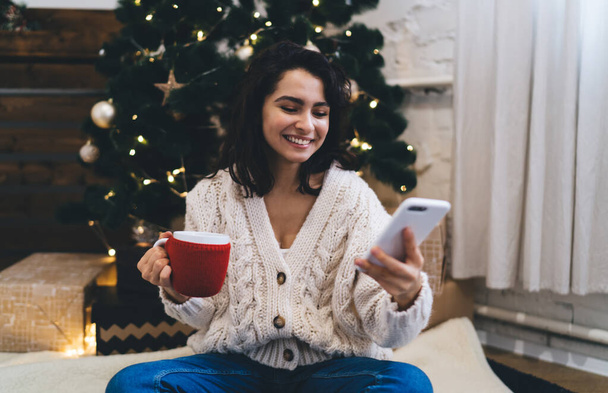 Young cheerful female in warm clothes with cup smiling and using smartphone near bright shiny decorated Christmas tree with presents - Fotoğraf, Görsel