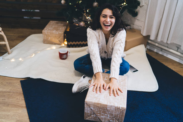From above of young positive female touching box with gift near Christmas tree with bright garland in comfortable contemporary lounge - Photo, Image