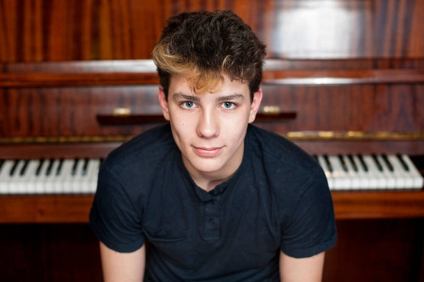 A cute teenager smiles against a piano background. He had a good mood because he played well at the concert. Horizontal. The boy has beautiful eyes and dark painted hair - Photo, image