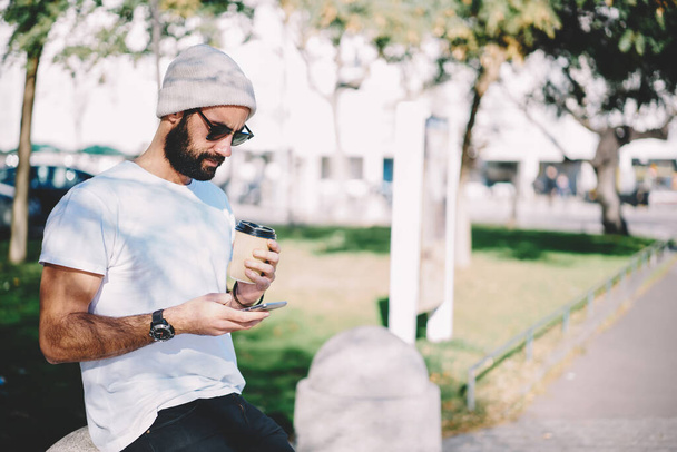 Middle Eastern male tourist in sunglasses reading web text while browsing page in social media networks, bearded hipster guy in winter hat browsing content and writing mail message via mobile app - 写真・画像