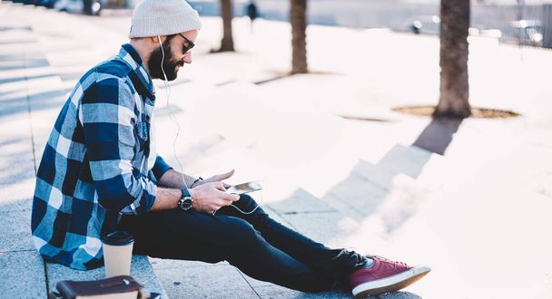 Middle Eastern male blogger connecting to 4g internet for installing media app on modern smartphone device using electronic headphones, Turkish hipster guy choosing music songs for listening - 写真・画像
