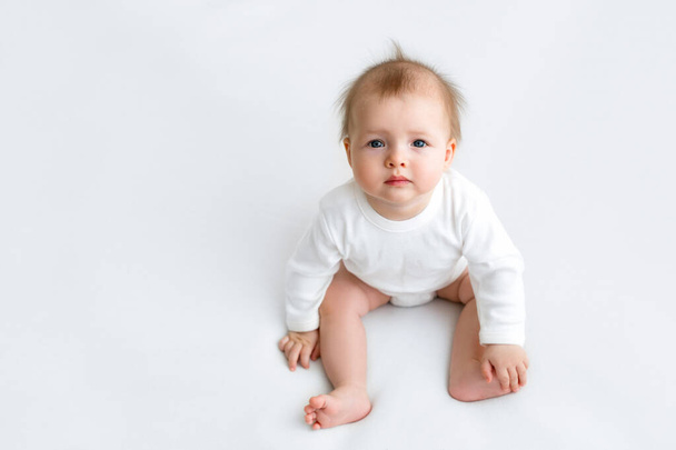 Βρέφος με προεξέχοντα μαλλιά κάθεται κοιτάζοντας την κάμερα. Αστεία μωρό σε λευκό φόντο σε ένα σώμα κοστούμι. Φροντίδα και φροντίδα των παιδιών από τη γέννηση. Το κορίτσι κρατάει το πόδι της για ισορροπία. - Φωτογραφία, εικόνα