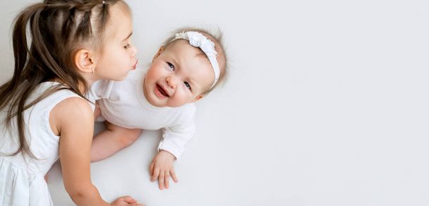Un bebé de seis meses solloza evitando los besos de su hermanita. Relación de los niños en una familia con una diferencia de edad. Chicas en ropa blanca en un fondo de estudio blanco con un espacio de copia - Foto, imagen