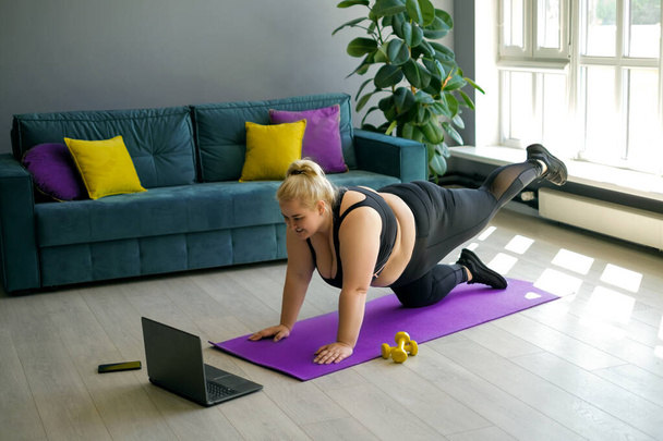 Curvy γυναίκα κάνει αθλήματα με έναν εκπαιδευτή σε απευθείας σύνδεση στο σπίτι στο σαλόνι, επαναλαμβάνοντας ασκήσεις από ένα βίντεο σε ένα φορητό υπολογιστή. Συνεδρία online προπόνηση. - Φωτογραφία, εικόνα