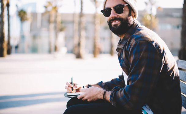 Portrait of trendy dressed male student with beard looking at camera while learning at urban bench in city, Middle Eastern writer in trendy sunglasses spending travel time for planning trip - Foto, immagini