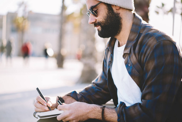 Talented Turkish male poet writing text ideas in copybook for creating articles, Middle Eastern hipster guy in sunglasses spending leisure daytime for note travel impression to personal diary - Photo, image