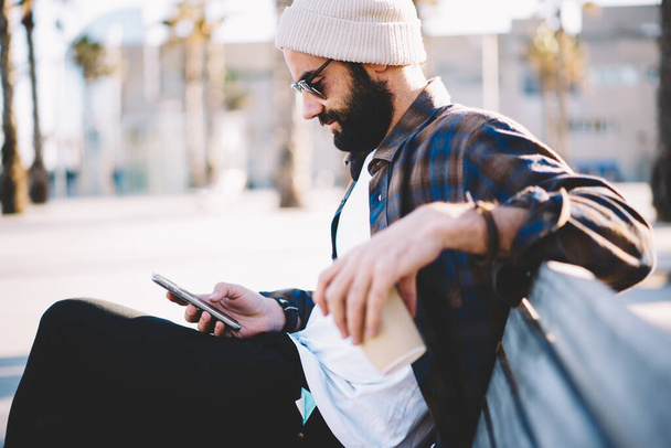 Middle Eastern male blogger in sunglasses and trendy hat checking received email message using 4g internet during leisure in city, hipster guy with coffee to go browsing wireless on smartphone - Φωτογραφία, εικόνα