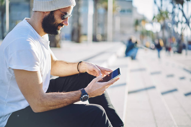 Millennial bearded man writing mail during smartphone chatting in social networks using 4g wireless on modern mobile gadget, Middle Eastern hipster guy reading travel publication resting in city - Foto, afbeelding