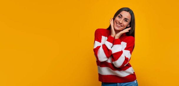 I'm so happy! Portrait of a smiling beautiful woman while she holds her palms on her cheeks and looking on camera on yellow background - Fotó, kép