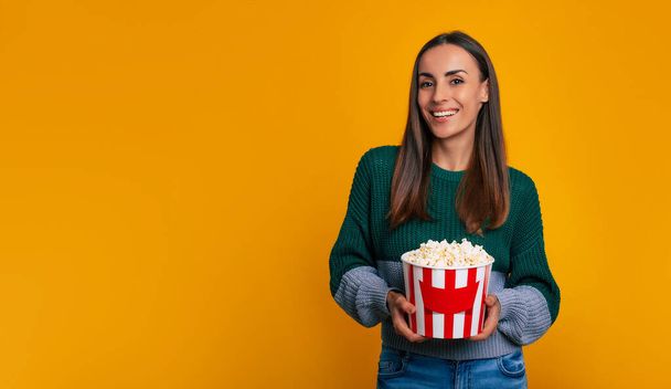 Session of blockbuster! Portrait of young gorgeous brunette girl while she holds big bucket of popcorn for watching movie in cinema and isolated on yellow background - Photo, image
