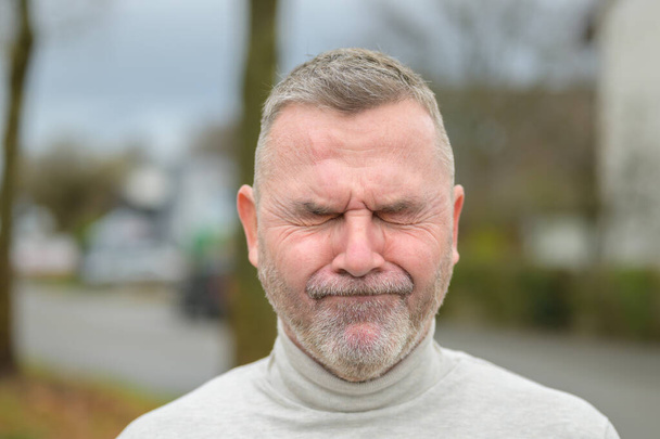 Embarrassed or anguished senior man with beard screwing up his face in a close up headshot in a quiet residential street - Foto, immagini
