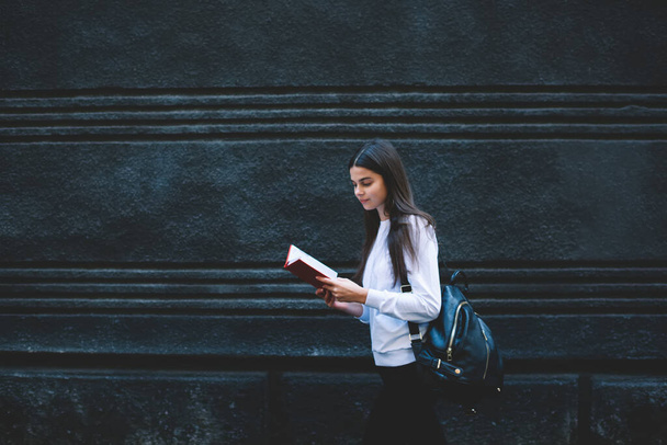 Side view of young dreamy female student wearing casual outfit and backpack standing near stone wall and reading interesting novel - Fotografie, Obrázek