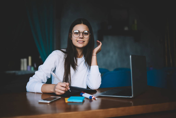 Smiling young brunette in eyeglasses wearing casual clothing sitting at wooden desk with portable computer and markers while writing in copybook and looking at camera - Photo, image
