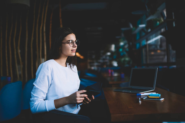 Side view of optimistic female remote worker with eyeglasses looking away while sitting at table with opened planner and working - Foto, immagini
