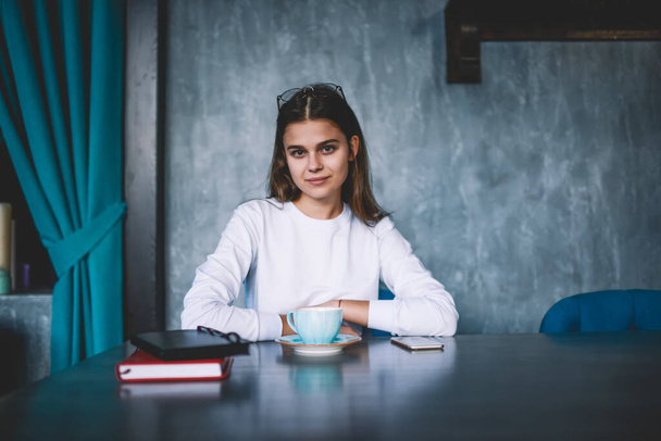 Young positive female student in casual wear sitting at table with cup of hot drink and books in cafeteria and looking at camera - Φωτογραφία, εικόνα