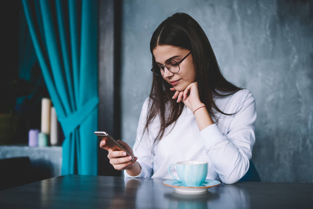 Pensive female in casual outfit with eyeglasses sitting at table with cup of tea leaning on hand and browsing mobile phone - Foto, Imagem