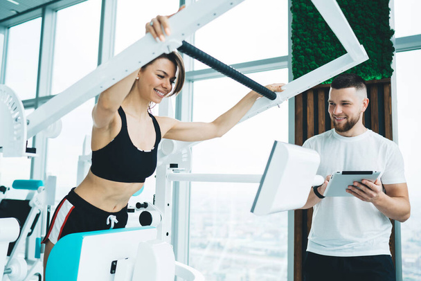Crop young cheerful female athlete in sportswear exercising on electronic equipment near window and glad instructor with tablet in gym - Foto, Imagen