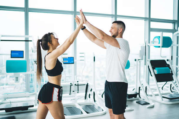 Side view of content sportswoman in active wear with smiling partner giving five against fitness machines while looking at each other - Photo, image