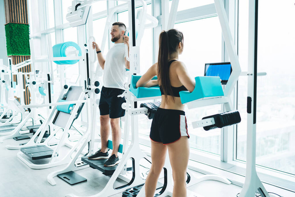 Athletic sportswoman with long hair exercising on fitness equipment near sportsman in activewear in modern spacious gym during intense training - Zdjęcie, obraz