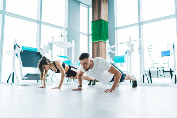 Full body of strong young sportsman and sportswoman in activewear training and doing push ups in spacious light large gym - Foto, Imagem