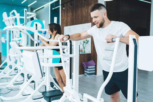 Strong bearded man in sportswear and wristwatch doing exercises on elliptical machine while training with coach in modern fitness center - Zdjęcie, obraz
