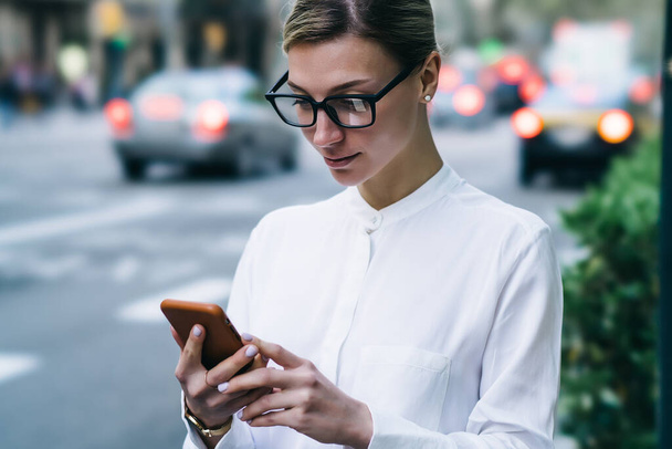 Crop pretty young female in smart casual outfit and eyeglasses looking at screen while standing by road and browsing smartphone - Фото, зображення