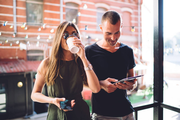 Half length of casual dressed hipster girl with modern mobile phone drinking tasty caffeine beverage while Caucasian boyfriend browsing web publication on digital tablet, millennial persons - Photo, Image