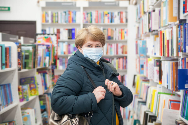 Felnőtt nő portréja orvosi maszkban a könyvesboltban, polcok közelében, tudományos irodalommal.  - Fotó, kép
