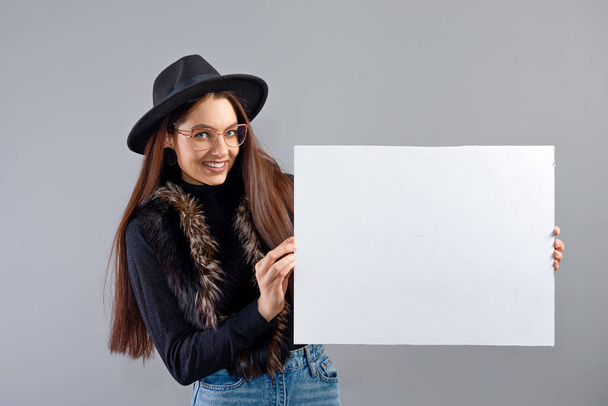 elegant young woman in glasses and hat showing blank board banner, isolated on gray, copy space - Valokuva, kuva
