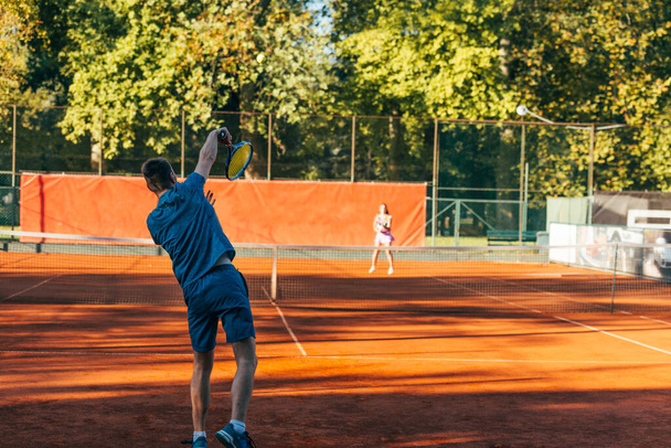 Visão traseira de um jogador de tênis pronto para servir em uma quadra de barro usando roupas esportivas azuis - Foto, Imagem