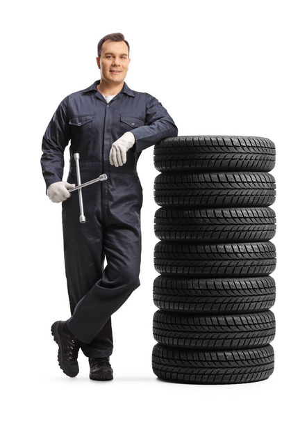 Auto mechanic worker in a uniform leaning on a pile of tires and holding lug wrench tool isolated on white background - Zdjęcie, obraz