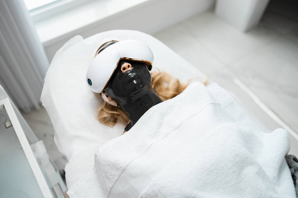 Blonde woman with a black alginate mask on her face and eye massage glasses lies on a couch of a beauty salon. Spa procedures and relaxation	 - Photo, Image